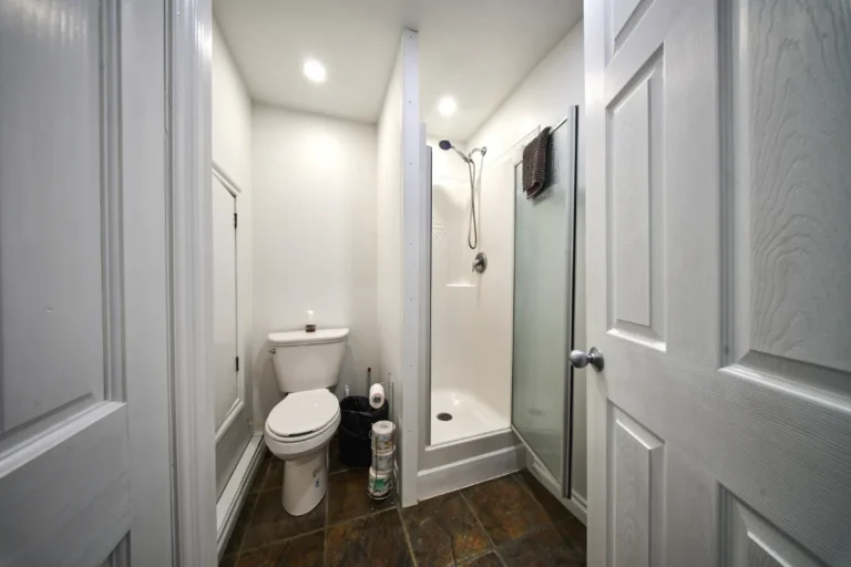 Narrow bathroom with a white toilet, a corner shower with a frosted glass door, and cleaning supplies on the floor next to the toilet.