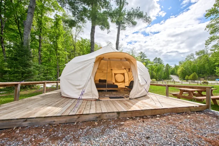 Beige canvas glamping tent pitched on a wooden deck in a forest campground, with chairs visible inside.