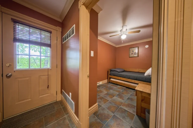 Small bedroom with a single bed along the far wall, ceiling fan, and a wooden nightstand in a warmly colored room near a doorway with a window showing greenery outside.