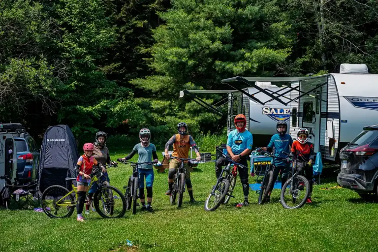 Group of cyclists with helmets on mountain bikes at a campground, with tents and a travel trailer in the background.