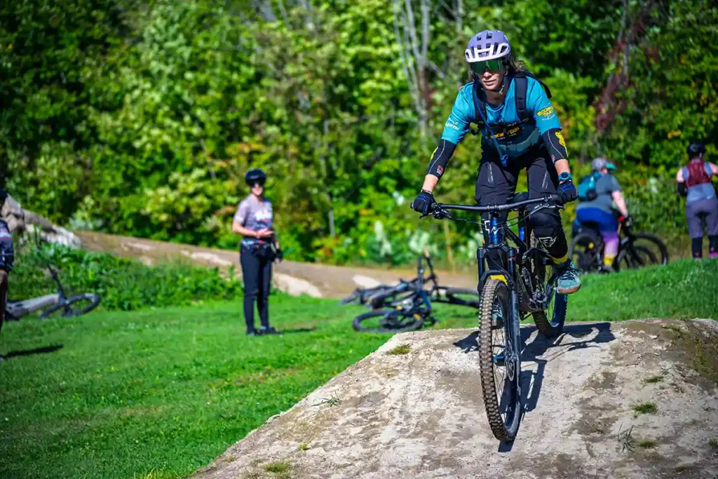 Cyclist wearing a blue jersey and helmet rides a mountain bike over a rocky ledge while spectators watch in a grassy park area.